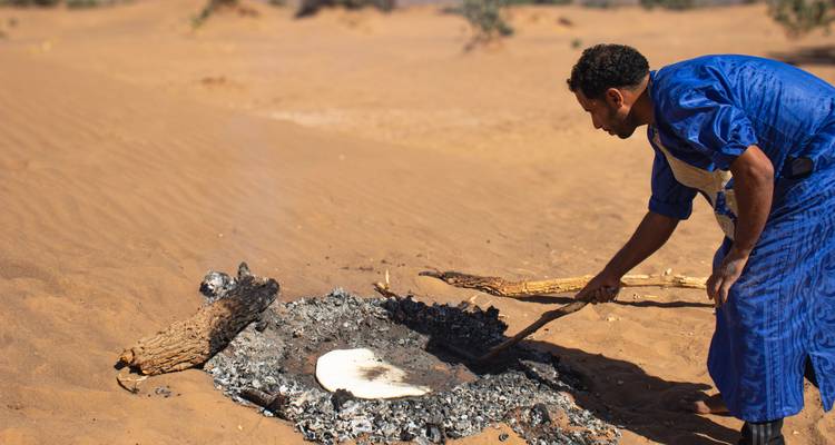 Local man bakes flatbread in hot desert sand and embers during a traditional cooking demonstration.