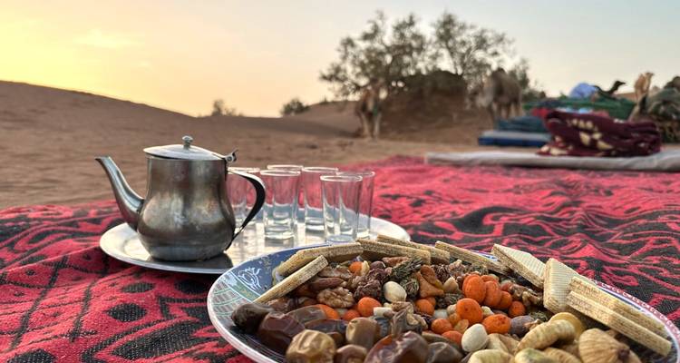 Plate of nuts, dates and snacks with tea set on patterned rug at desert camp as camels stand in background at dusk.