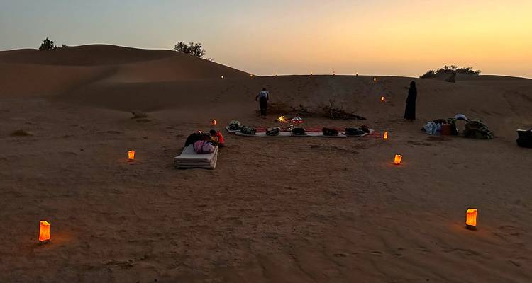 Small desert camp with mattresses and lanterns laid out under twilight sky between sand dunes.