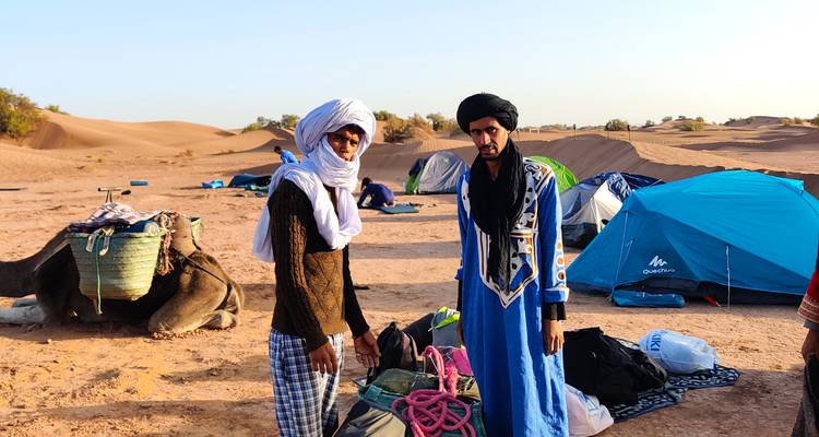 Two men in traditional attire stand by camping gear and tents in a desert expedition setting.