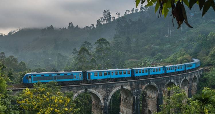 A bright blue train curves across the historic Nine-Arch Bridge amid misty green hills.