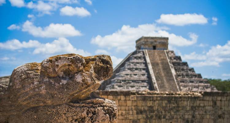 Stone serpent head foregrounds a creative view of the Chichen Itza pyramid against blue sky.
