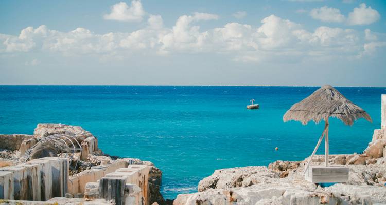 Clear teal water laps against rocky shore with a single palapa umbrella and boat offshore at Isla Mujeres.