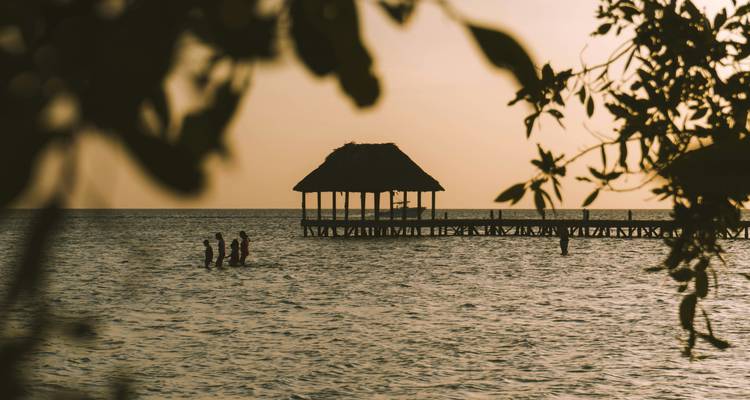 Silhouettes of people standing in shallow water near a wooden pier hut at dusk on Holbox Island.