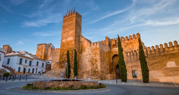 Muros y torres de castillo de arenisca medieval dominando una plaza tranquila con una pequeña fuente.
