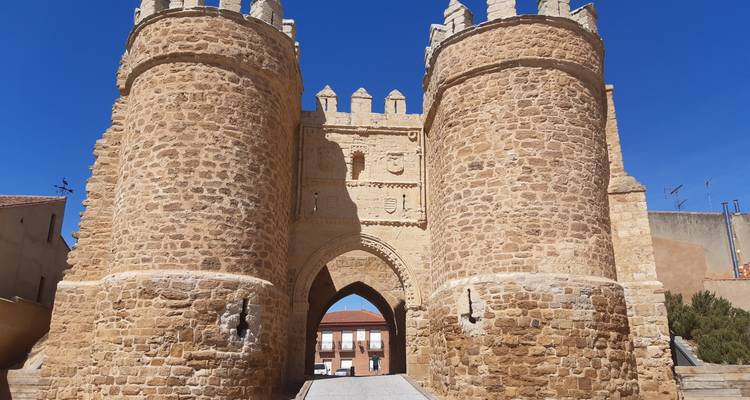 Impresionante puerta de piedra con dos torres gemelas y un pasaje arqueado que lleva a una plaza soleada del pueblo.