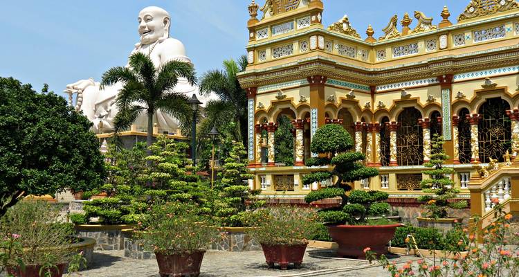 Ornate Vinh Trang Pagoda complex with manicured bonsai and giant laughing Buddha statue.