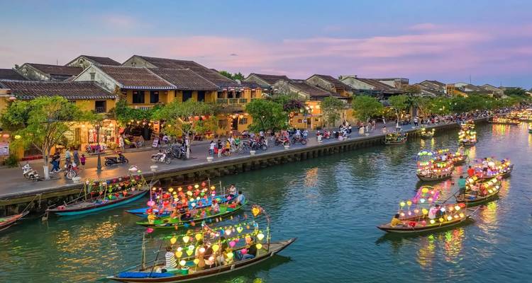 Lantern-lit boats drift along Hoi An’s waterfront at colorful sunset with ancient houses behind.