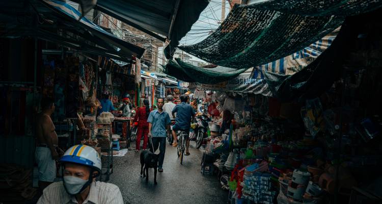 A bustling, narrow market alley packed with vendors, cyclists, shoppers, and hanging tarps in low light.