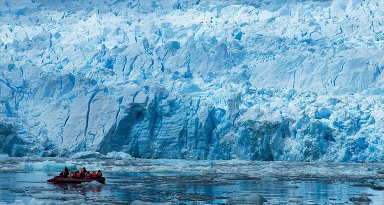 Des visiteurs dans un petit radeau dérivent le long d'une imposante paroi de glacier bleu tandis que des glaçons flottent autour d'eux.