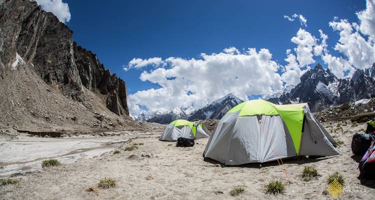 Row of expedition tents pitched on a sandy moraine beneath towering snow-capped Karakoram peaks.