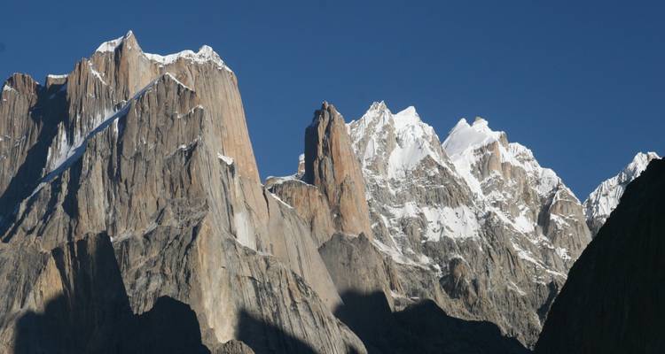Dramatic granite spires and snow-capped peaks of the Trango Towers against a deep blue sky.
