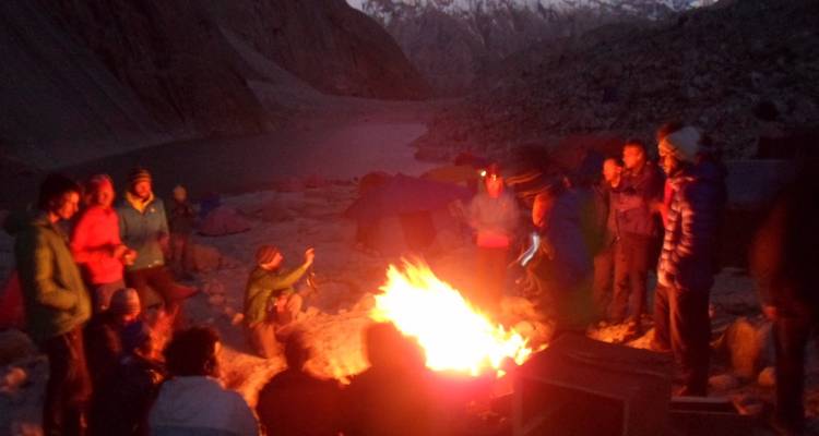 Trekking group gathered around a bright campfire at dusk beside tents and a mountain lake.