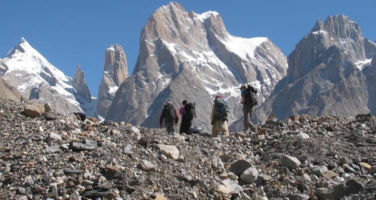 Trekking party traverses rocky glacial debris with jagged snow-covered towers looming overhead.