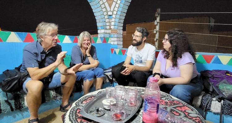 Les voyageurs conversent autour d'une table basse garnie de verres à thé vides à l'intérieur d'une chambre nubienne aux couleurs vives, la nuit.
