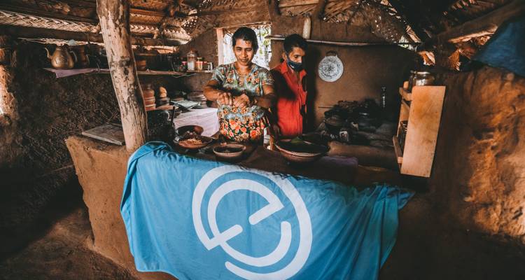 Two women prepare traditional dishes in a rustic clay kitchen draped with a blue travel banner.