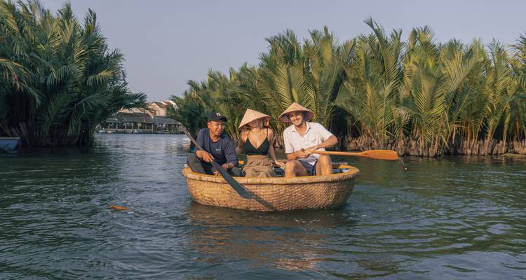 Three travellers paddle a round bamboo basket boat through lush nipa palms on a calm river.