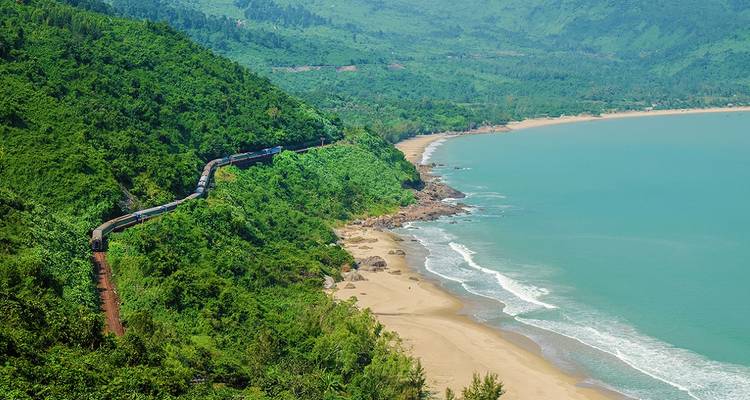 A coastal train curves along the lush green Hai Van Pass overlooking a turquoise bay and sandy beach.