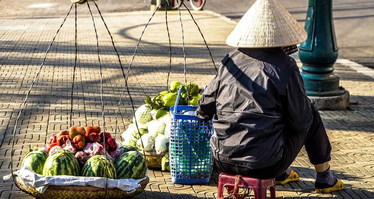 Un vendedor callejero vietnamita con un sombrero cónico se sienta junto a canastas de productos frescos equilibradas en varas de bambú.