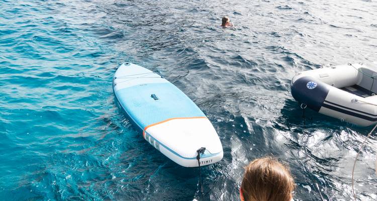 Inflatable paddleboard in clear blue water with swimmer nearby and dinghy tied up.