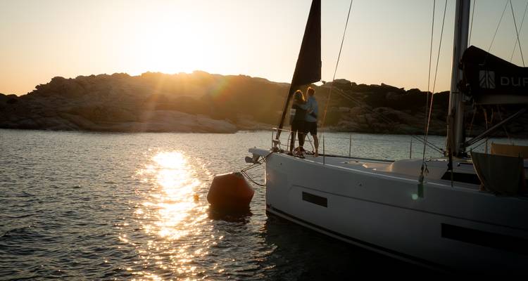 Couple standing on the bow of a sailboat enjoying a glowing sunset over rocky islands