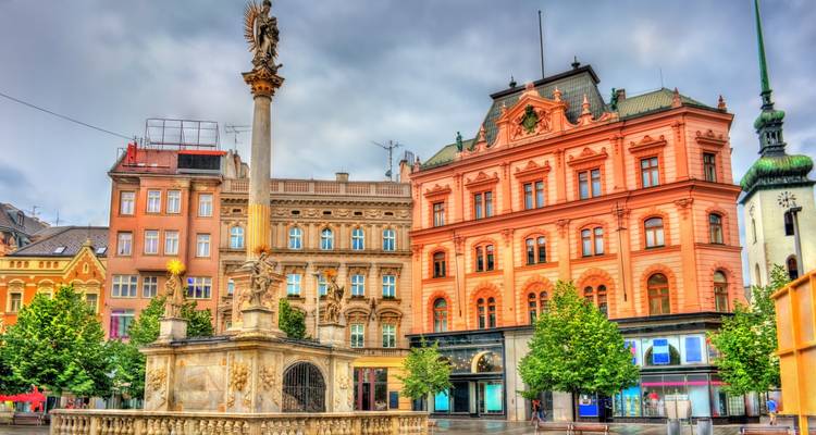 Place historique colorée avec fontaine ornementée, façades pastel et arbres feuillus sous un ciel gris.
