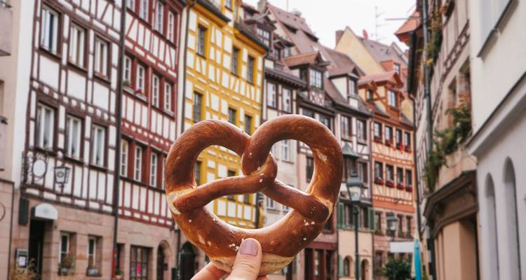 Primer plano de un pretzel tradicional sostenido frente a casas con entramado de madera de colores en Núremberg.