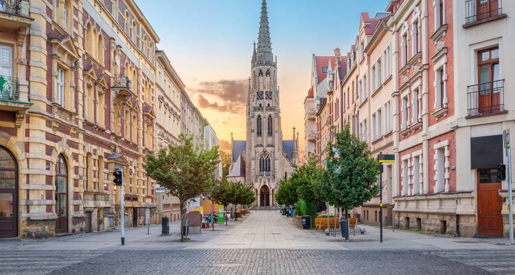 Rue symétrique de la ville de Wroclaw menant à une haute flèche d'église néo-gothique, encadrée par des façades d'immeubles d'appartements ornées au coucher du soleil.