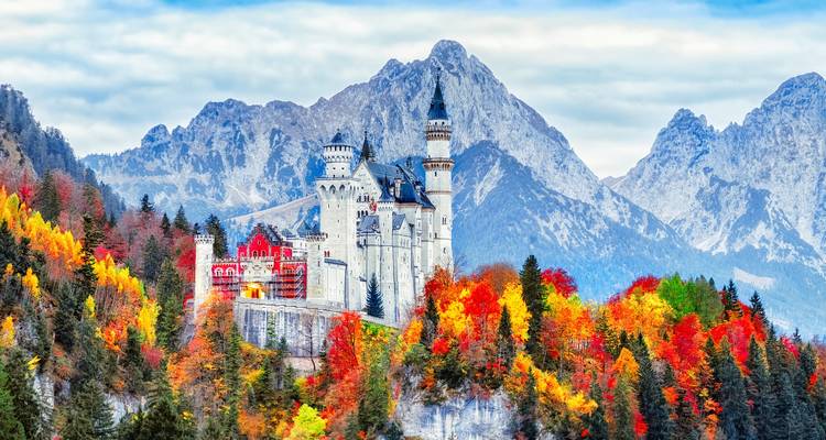 Sprookjesachtig Neuschwanstein Castle omgeven door kleurrijk herfstwoud met dramatische Alpentoppen op de achtergrond.
