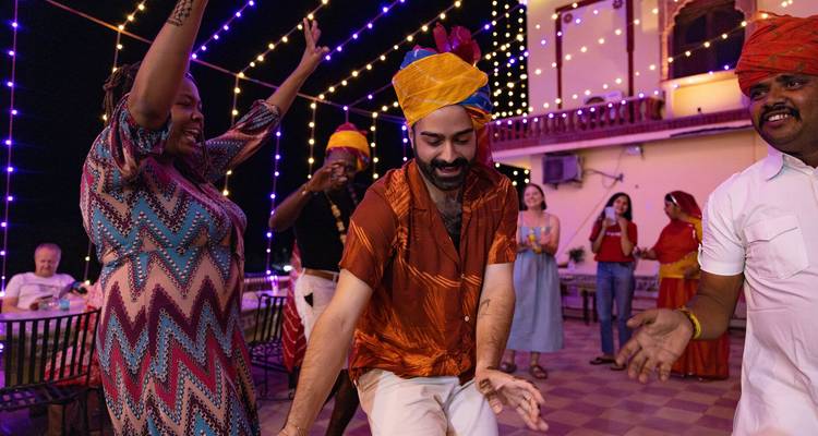 Travelers wearing colorful turbans dance joyfully under strings of festival lights at night.