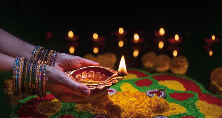 Hands adorned with colorful bangles hold a lit clay lamp above a vibrant rangoli design.