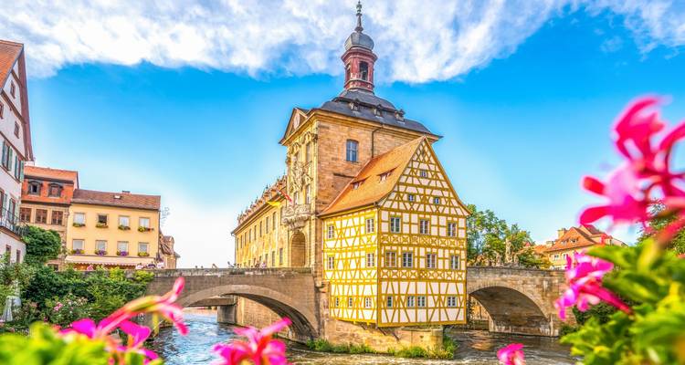 Vue éclatante de l'Hôtel de Ville ancien de Bamberg perché sur un pont de pierre encadré par des fleurs rose vif et un ciel bleu.