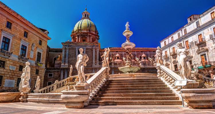 Les statues de marbre et les grands escaliers de la Fontana Pretoria encadrés par les bâtiments historiques de Palerme sous un ciel bleu profond.