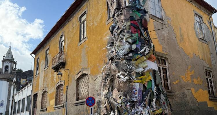 Ángulo amplio de una enorme escultura de gato hecha con materiales reciclados, con un edificio amarillo antiguo y el cielo azul de fondo.