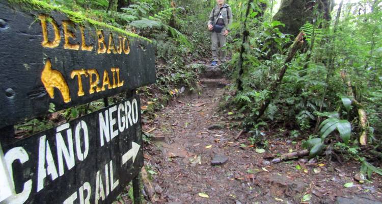 Hiker stands on muddy jungle trail beside wooden sign for Caño Negro Trail
