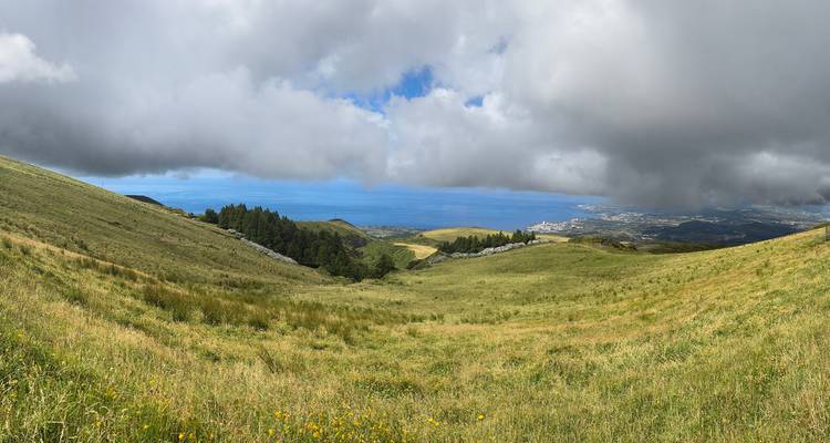 Valle amplio de praderas verdes que lleva al Océano Atlántico bajo nubes dramáticas con cielo azul que se asoma por entre ellas.