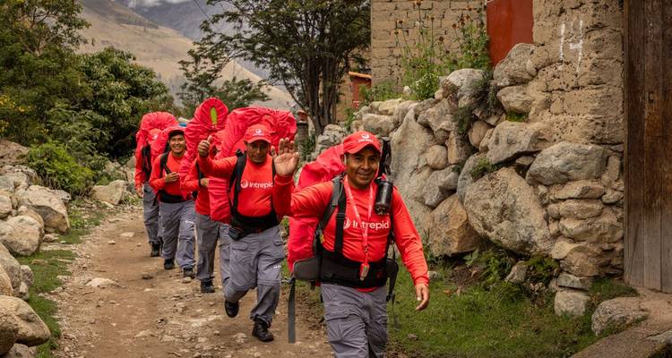 Des porteurs souriants vêtus d'uniformes rouges trottinent sur un sentier en pierre, portant de grands sacs à dos entre les villages de montagne.