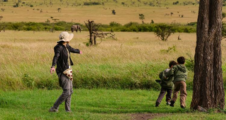 Un adulte et deux enfants marchant joyeusement à travers une savane herbeuse avec un éléphant visible au loin
