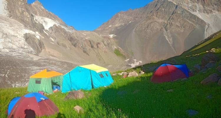 Brightly colored camping tents set in a lush alpine meadow beneath steep rocky peaks and glaciers.