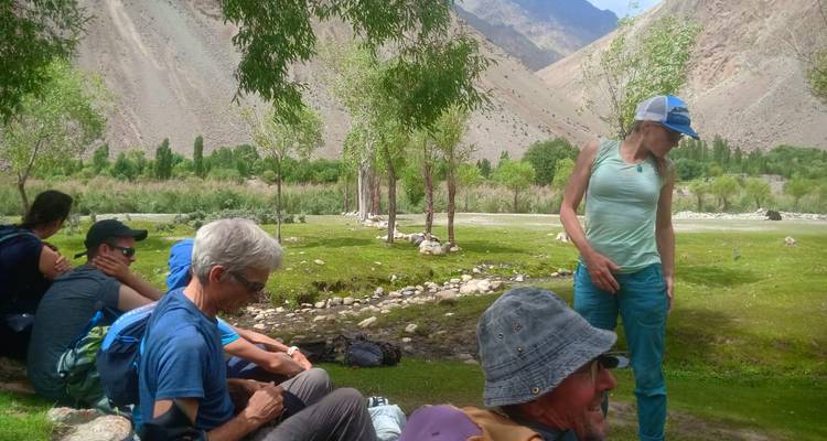 Trekking party rests in a shady riverside grove backed by arid mountains.