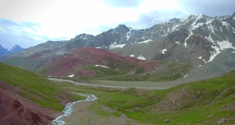 Wide alpine valley with red and green slopes, snow-dusted peaks and a winding stream.