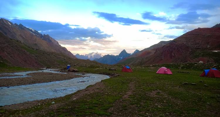 Tents pitched beside a winding river at dusk with distant jagged peaks silhouetted against sunset.