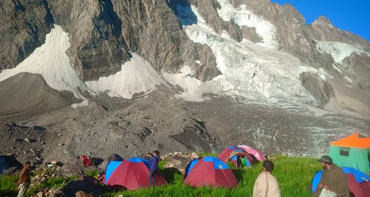 Cluster of tents on bright grass below a dramatic hanging glacier and dark rock walls.