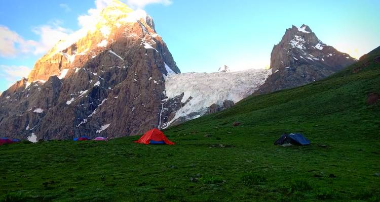 Red and blue tents on a green alpine meadow beneath sun-lit rocky spires and glacier remnants.