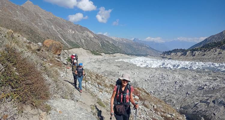 Line of hikers traversing a rocky trail beside a vast grey glacier under blue skies.