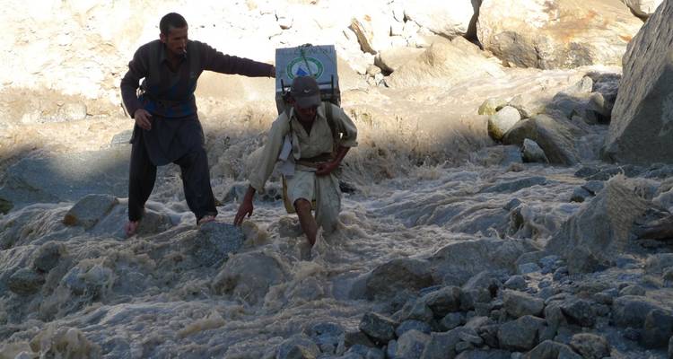 Two porters struggle across a raging melt-water stream carrying heavy loads.
