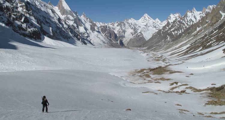 Lone trekker traverses a vast snow-covered glacier valley surrounded by spired peaks.