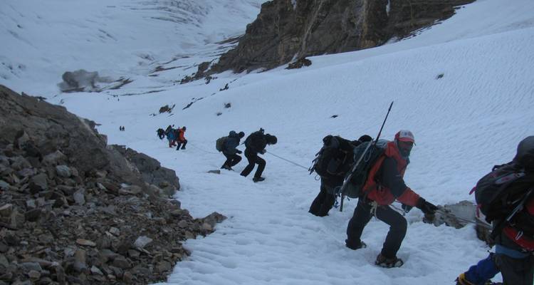 Climbers ascend a steep snow slope roped together against craggy mountain backdrops.