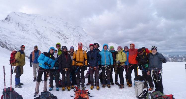 Expedition team in bulky climbing gear poses together on a snowy plateau below a massive peak.