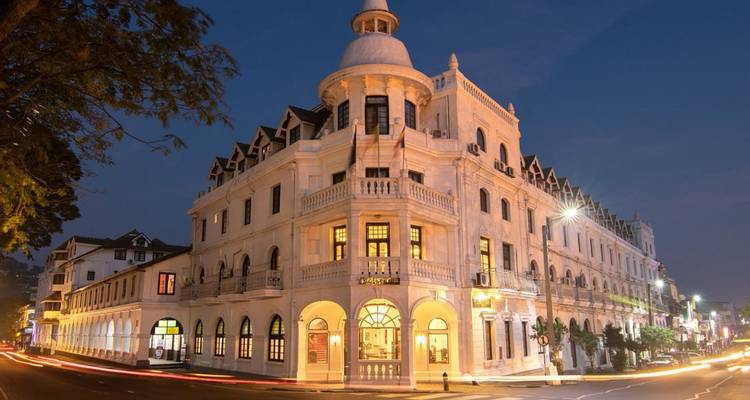 Elegant koloniaal hotel verlicht tijdens het blauwe uurtje op een rustige straathoek met lichtsporen van voorbijrijdend verkeer.